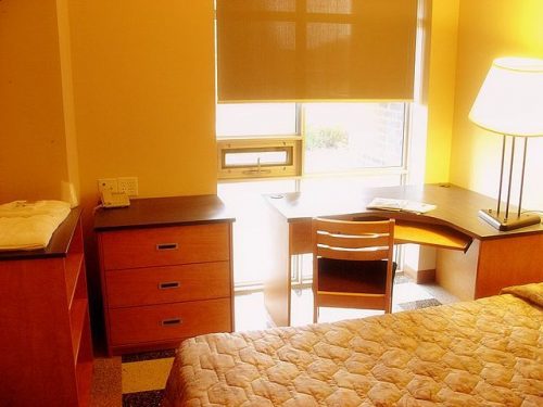 Photo of a dorm room with a desk, mattree, lamp